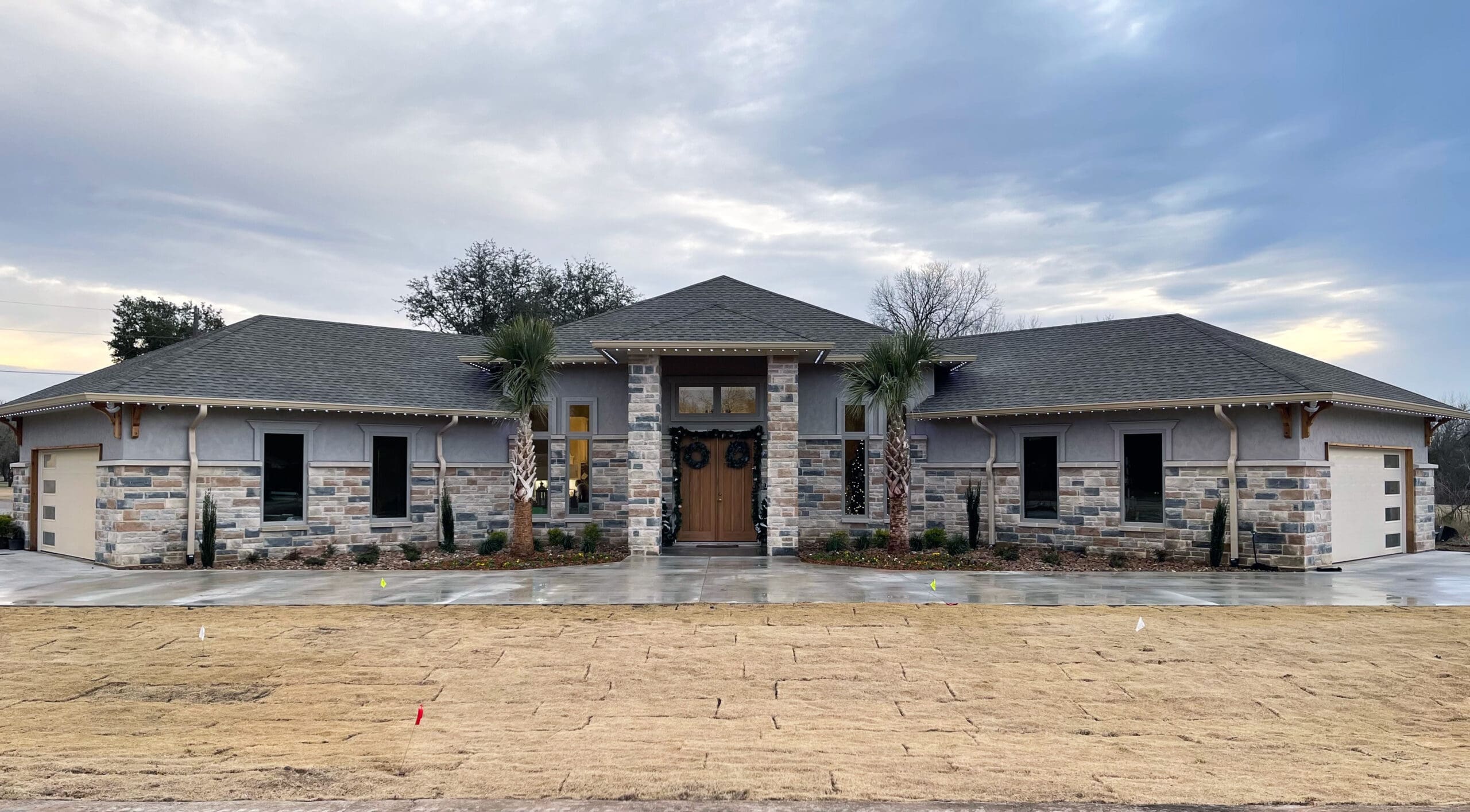 Wide view of a spacious stone ranch-style home with a symmetrical entrance, gray roof, driveway, sparse landscaping, and an overcast sky.