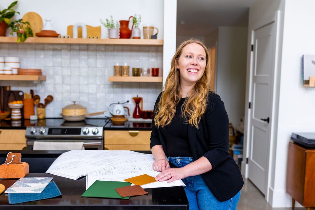Founder Carly Brotherson smiling while standing at a kitchen island with architectural plans and colored samples.